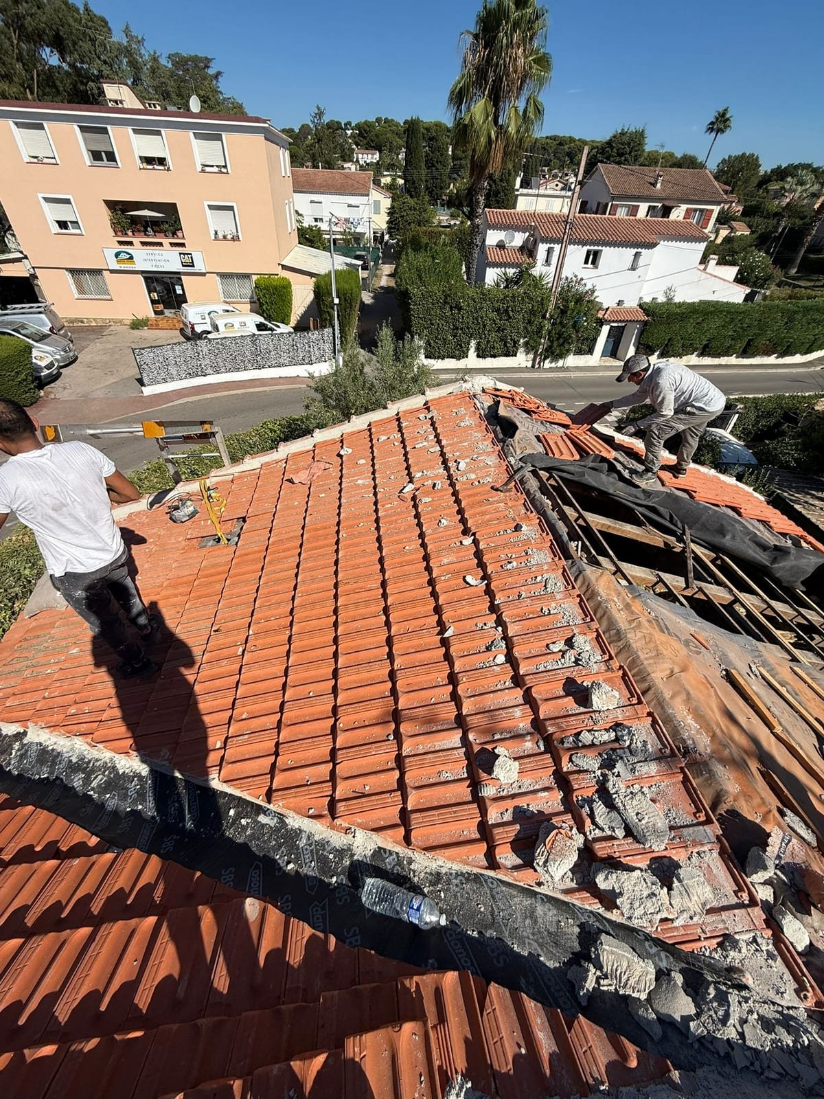 Deux couvreurs sur une toiture en cours de dépose, tuiles retirées et gravats de mortier sur le versant