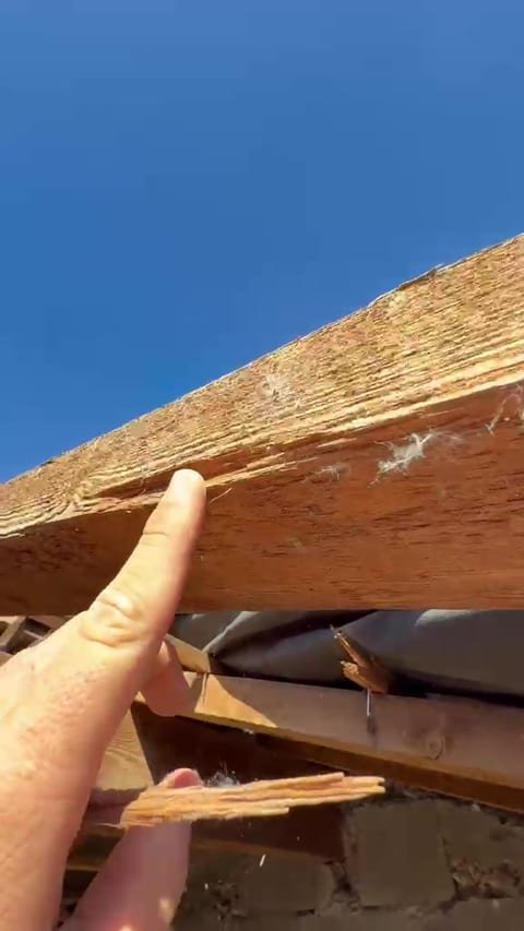 Plan rapproché sur une pièce de bois de toiture fendue et éclatée, désignée du doigt, ciel bleu en arrière-plan